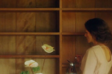 Woman stands inside a house with all dark oak wood interiors. Beside her is a vase with orchids