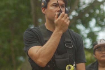 Bradley Tangonan wears camera gear and a headset, talking into a walkie-talkie to communicate with the rest of his film crew on site