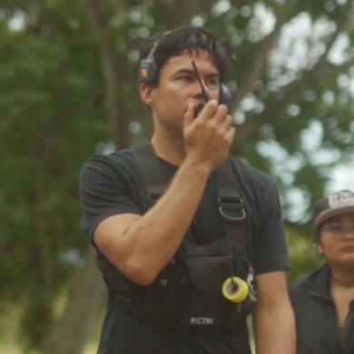 Bradley Tangonan wears camera gear and a headset, talking into a walkie-talkie to communicate with the rest of his film crew on site