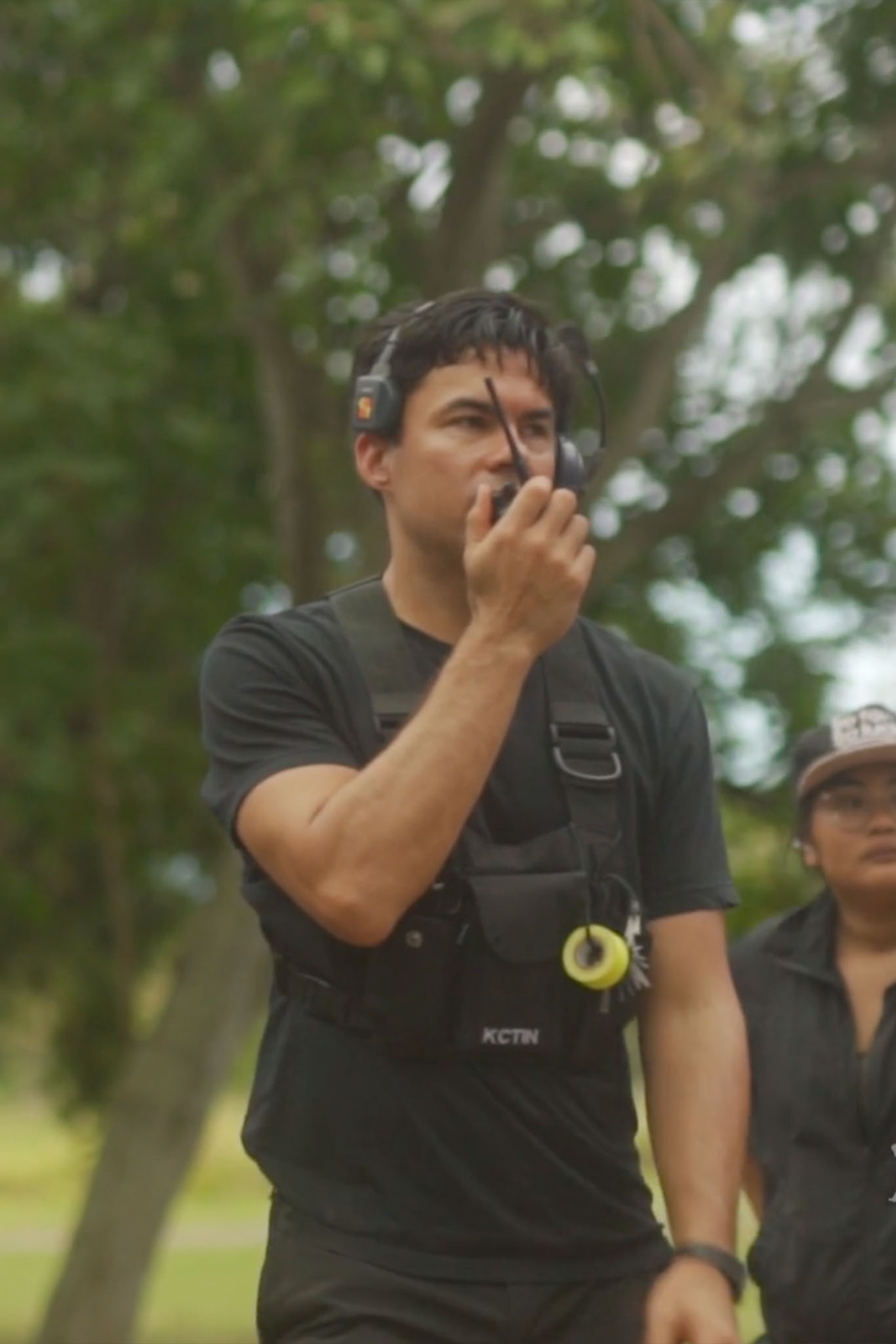 Bradley Tangonan wears camera gear and a headset, talking into a walkie-talkie to communicate with the rest of his film crew on site