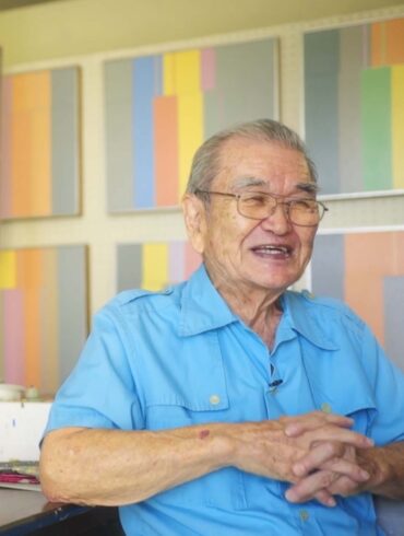 Harry Tsuchidana sits down in an interview, wearing blue, smiling with his color-block work in canvas behind him.