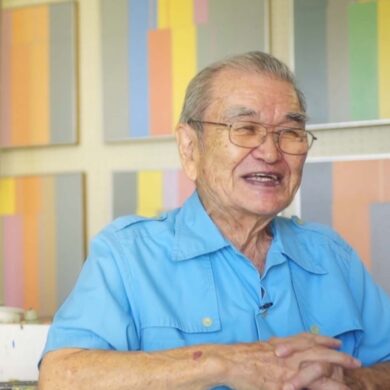 Harry Tsuchidana sits down in an interview, wearing blue, smiling with his color-block work in canvas behind him.