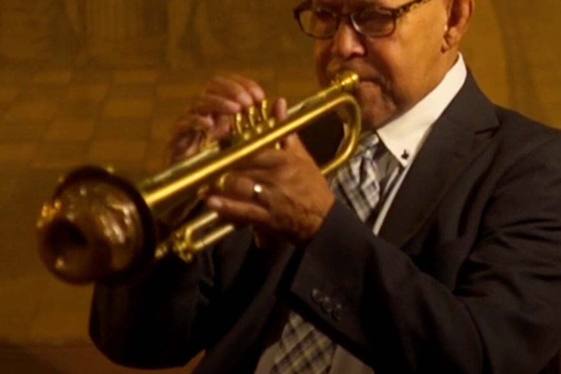 Trumpeter Eddie Henderson sits in a black and white suit, playing his gold trumpet