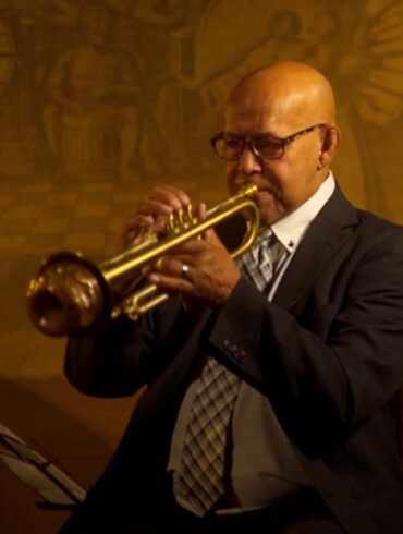 Trumpeter Eddie Henderson sits in a black and white suit, playing his gold trumpet