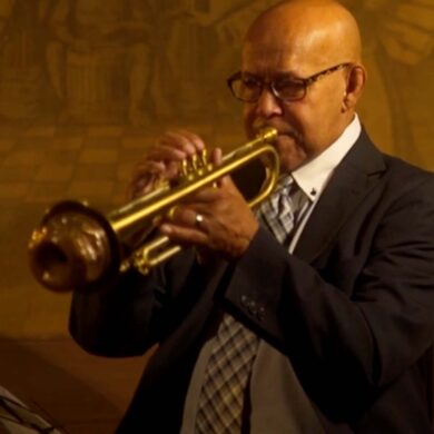 Trumpeter Eddie Henderson sits in a black and white suit, playing his gold trumpet