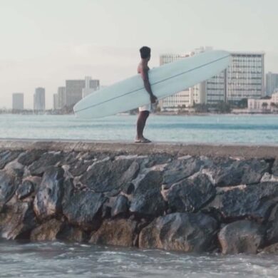 Arthur “Toots” Anchinges, stands at the edge of a pile of rocks by the beach shore -- looking out, holding his white surf board