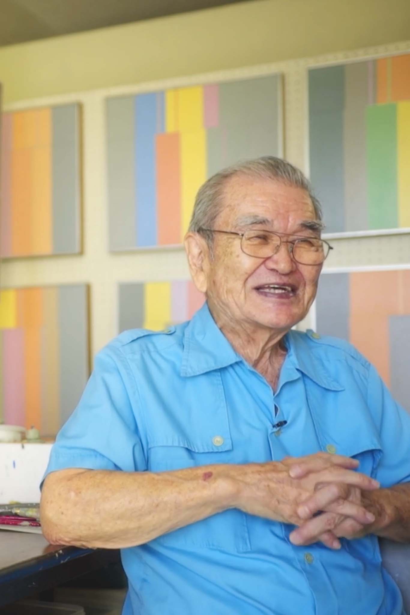 Harry Tsuchidana sits down in an interview, wearing blue, smiling with his color-block work in canvas behind him.