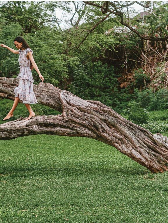 A person in a white floral dress walking on a large fallen tree trunk in a lush green park setting with trees and foliage in the background.