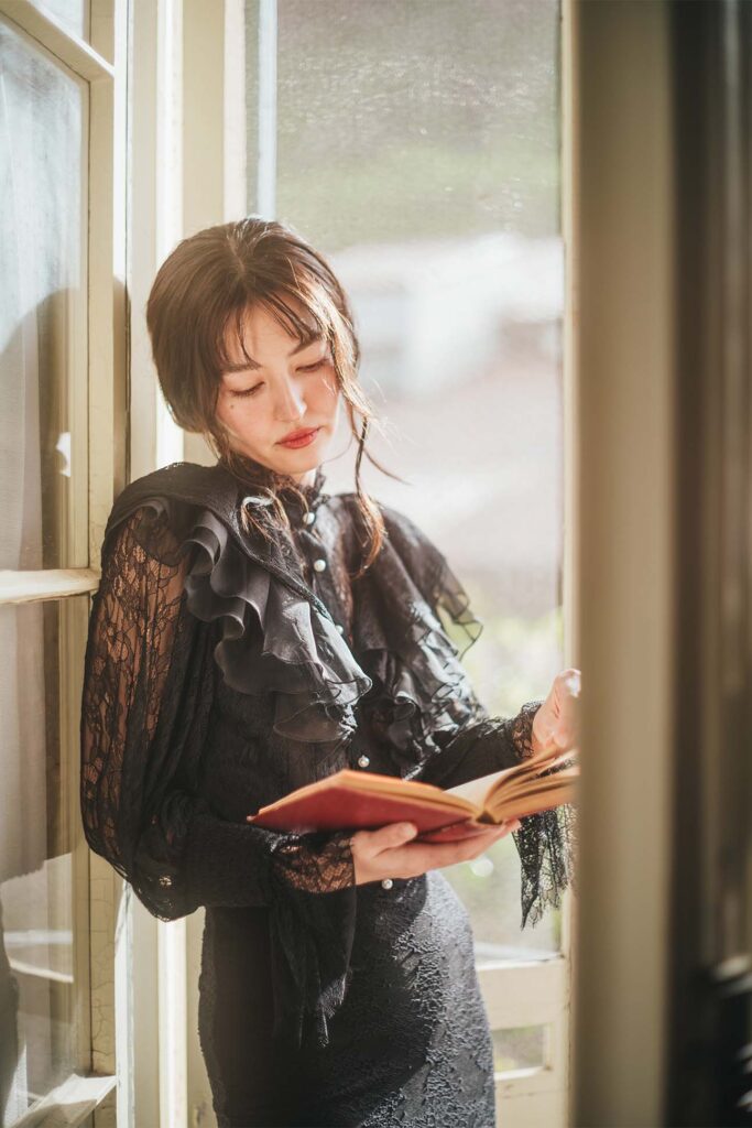 A person in a black lace dress standing by a window reading a book bathed in soft natural light.
