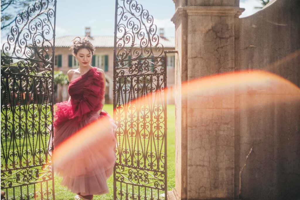 A person in a vibrant red ruffled dress stands behind an ornate wrought-iron gate, with a classical building and greenery in the background, all under bright sunlight with a lens flare effect.