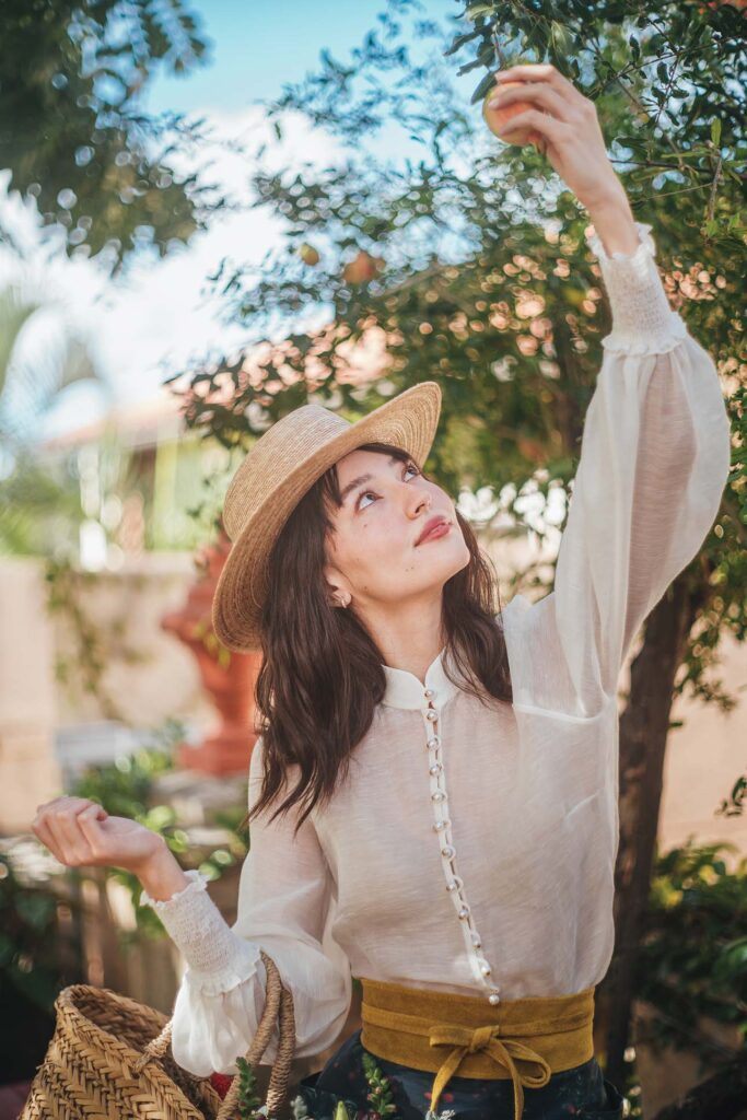 A person in a straw hat and a sheer white blouse with puffed sleeves, wearing a golden belt and a floral skirt, standing in a sunny garden, reaching up to pluck a fruit from a tree, with a wicker basket on one arm.