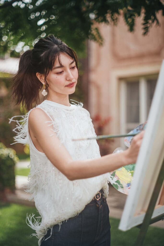 A person in a white feathered sleeveless top and jeans stands in front of an easel, holding a palette with various colors of paint and a paintbrush, seemingly in the act of painting. The setting appears to be an outdoor area with greenery and a building in the background.