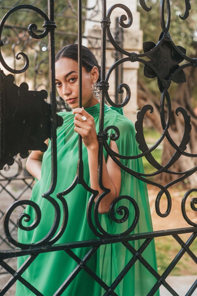 A person in a vibrant green dress with pleat detail stands behind a black ornate wrought iron gate, holding onto the bars with manicured hands adorned with a large ring and ornate earrings.