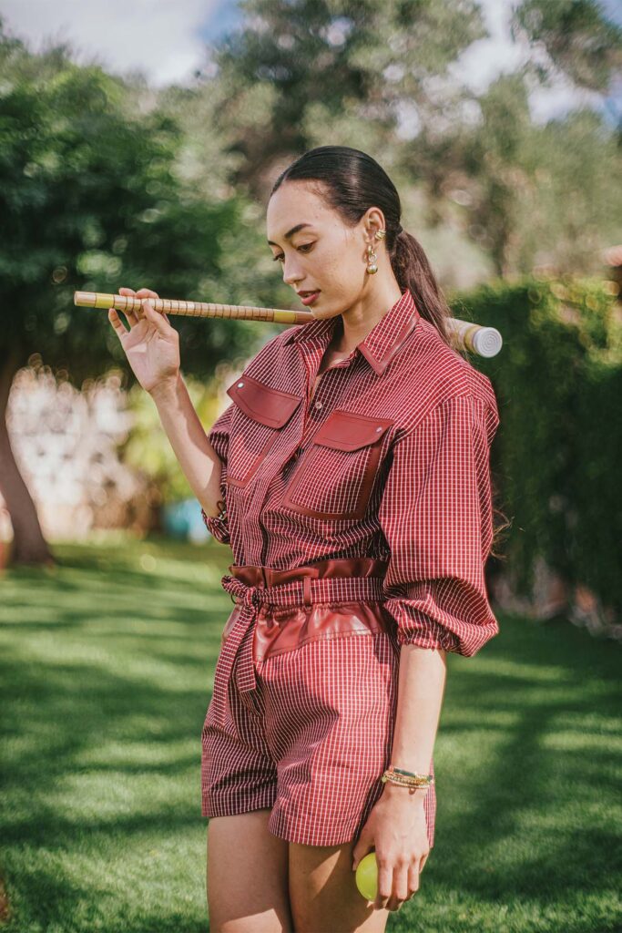 A person is holding a flute to their side while wearing a red and white checked shirt with rolled-up sleeves and matching shorts, accented with brown leather details. They are accessorized with large hoop earrings and multiple bracelets, holding a lemon in one hand against a background of lush greenery.