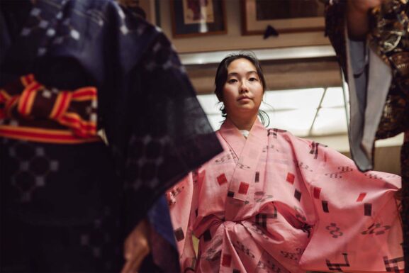A person wearing a pink kimono with square and floral patterns sitting indoors, with another individual in a dark kimono and patterned obi belt partially visible on the left.