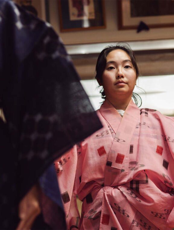 A person wearing a pink kimono with square and floral patterns sitting indoors, with another individual in a dark kimono and patterned obi belt partially visible on the left.