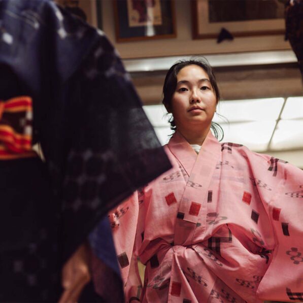A person wearing a pink kimono with square and floral patterns sitting indoors, with another individual in a dark kimono and patterned obi belt partially visible on the left.