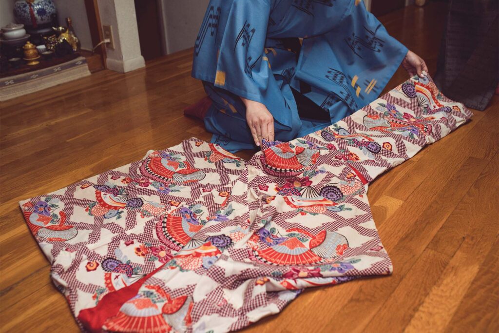 Person wearing a traditional blue kimono kneeling on wooden floor and folding a vibrant red and white patterned fabric with fan designs.