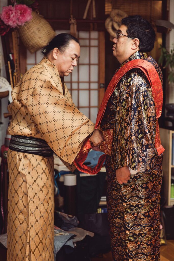 Two individuals standing side by side wearing traditional Japanese kimono, one in a beige kimono with a black obi belt and the other in an ornate black kimono with a red and gold obi, holding hands in front of a room with Japanese decor.