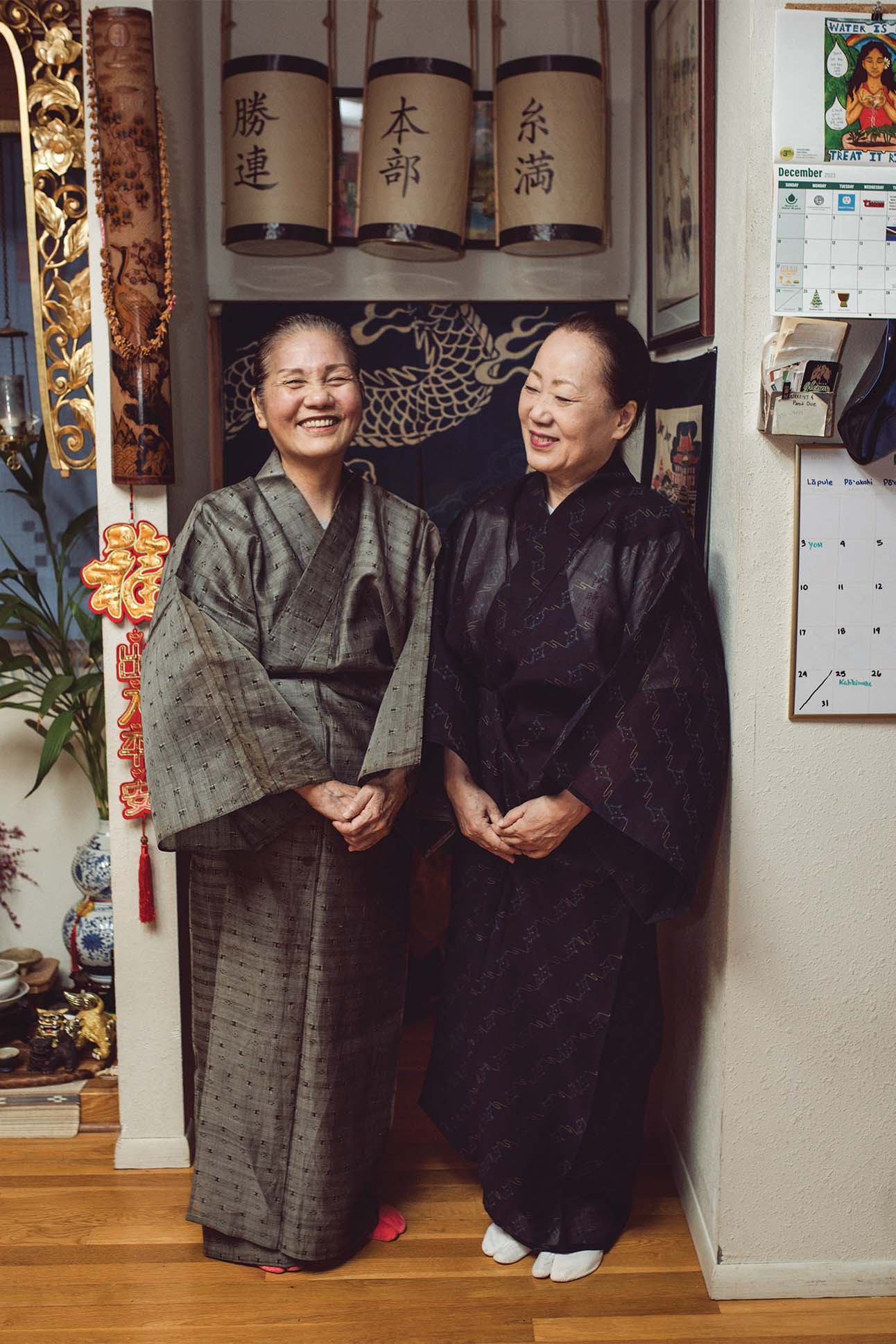 Two individuals wearing traditional Japanese kimonos standing in a room with cultural decorations, including hanging scrolls with Japanese writing, a wall calendar, and a red Chinese knot ornament. The room also features various ornamental objects and a peek into an adjacent area with a bamboo scroll painting.