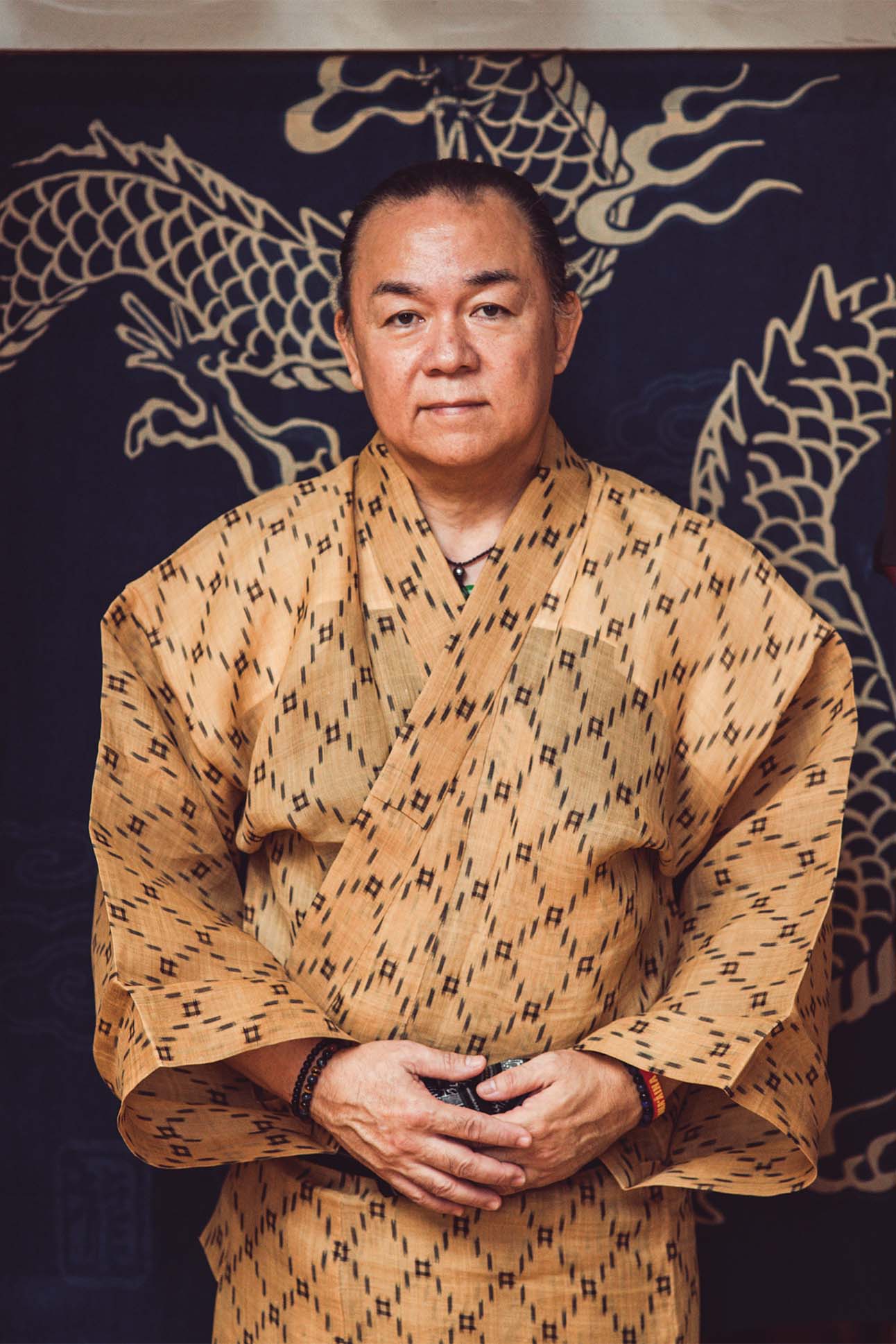 A person wearing a traditional patterned kimono with folded arms in front of a dark backdrop featuring a large white dragon design.