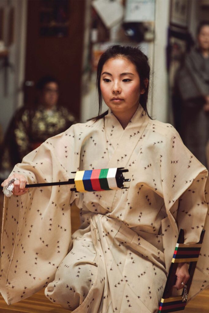 A person wearing a traditional cream-colored patterned kimono with wide sleeves holds a Kendo Shinai bamboo sword with multicolored handle tape.
