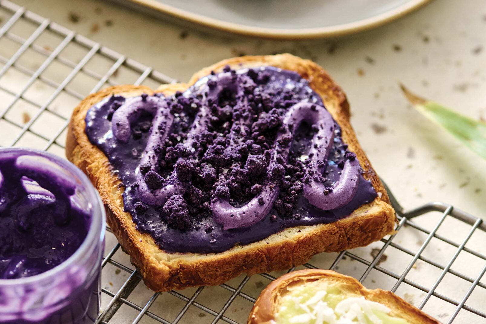 A piece of toast with vibrant purple yam spread and crumbled toppings on a cooling rack with a jar of spread and plate in the background.