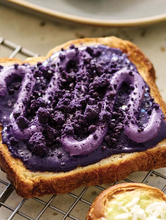 A piece of toast with vibrant purple yam spread and crumbled toppings on a cooling rack with a jar of spread and plate in the background.