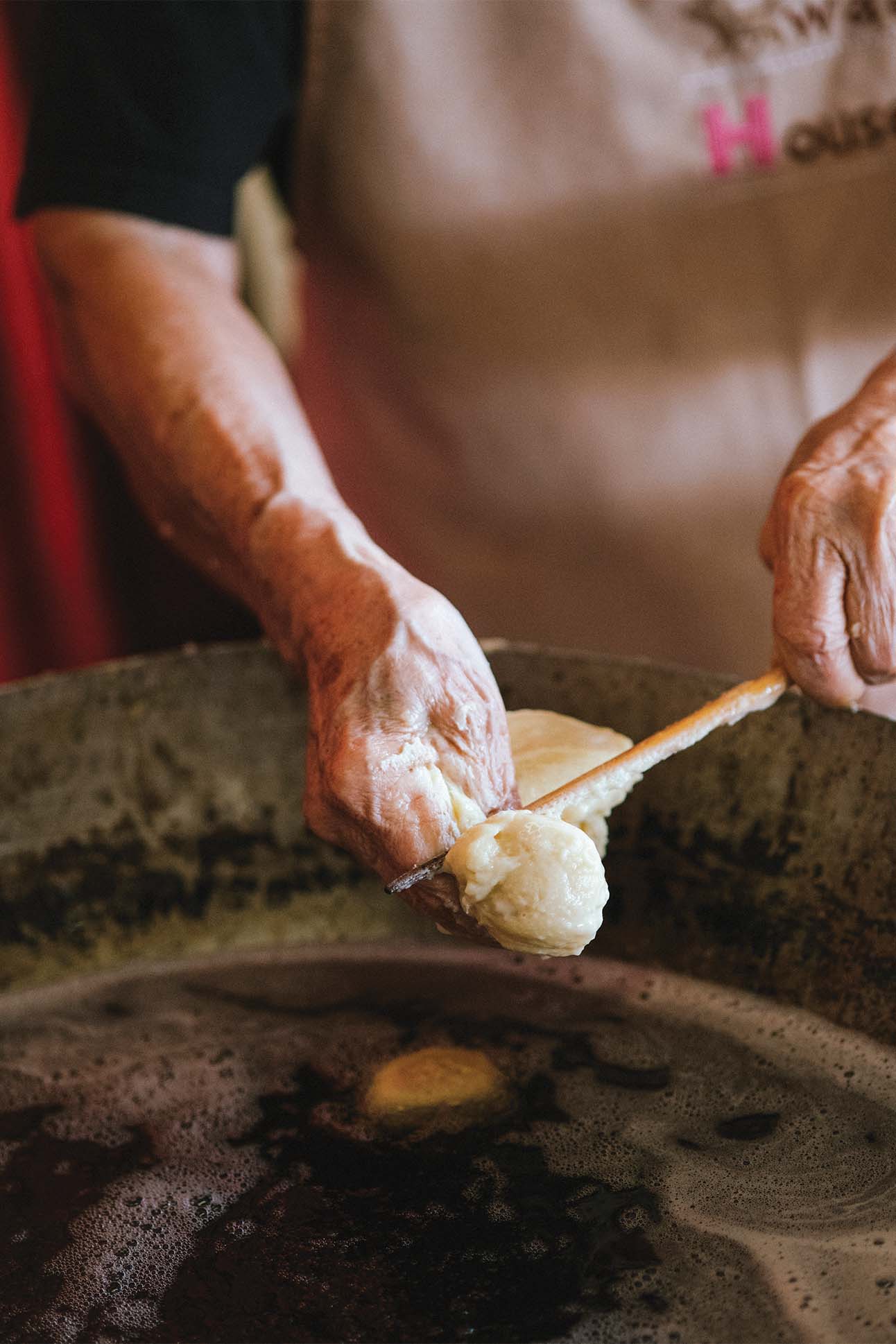An elderly person's hands, wearing an apron, scooping homemade butter from a large wooden bowl with a flat utensil.