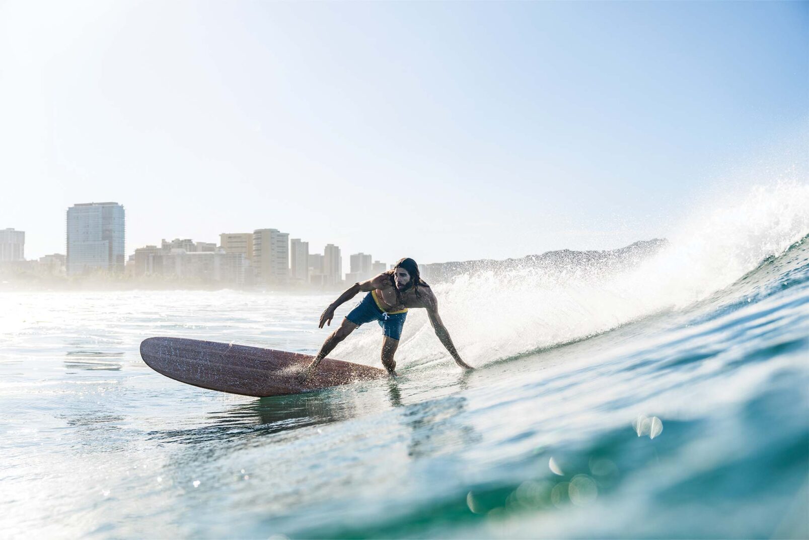 A surfer, wearing a black top and blue shorts, skillfully riding a wave on a longboard with a clear blue sky and a city skyline in the background.