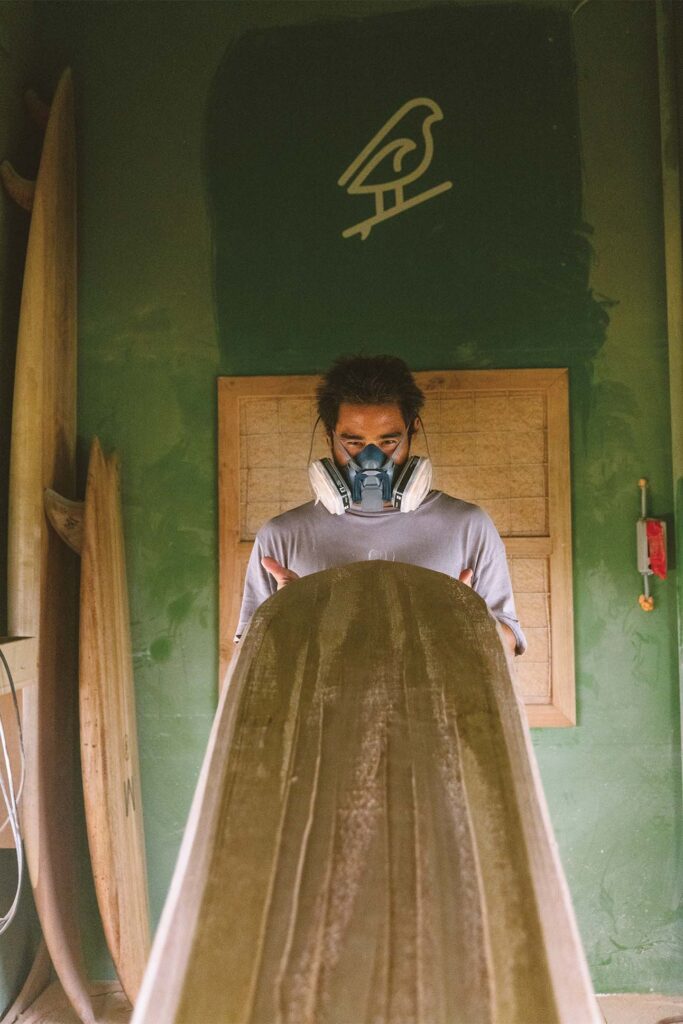 A person in a gray sweatshirt holding a large wooden surfboard in a room with green walls and a surfboard-shaped logo. Multiple surfboards are propped against the wall.