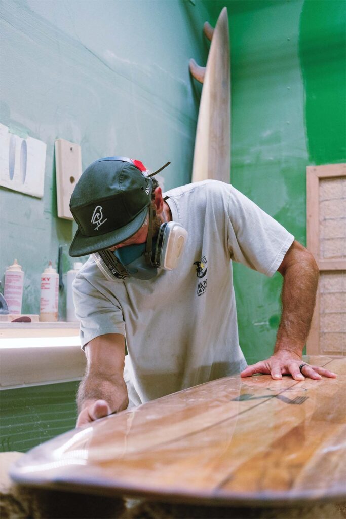 Man wearing a protective mask and hat sanding a wooden surfboard in a workshop.