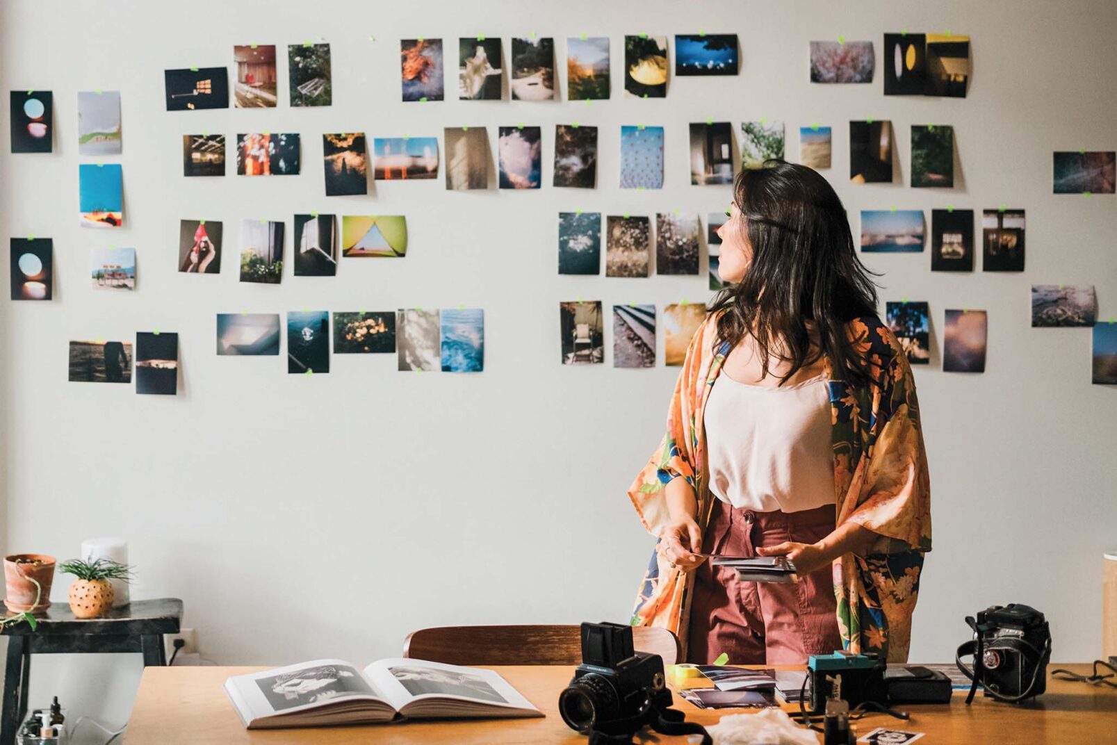 A person stands in front of a wall adorned with a collage of assorted photographs, facing away from the camera. They are surrounded by a creative workspace that includes photography equipment, an open book, a potted plant, and various personal items on a wooden table.
