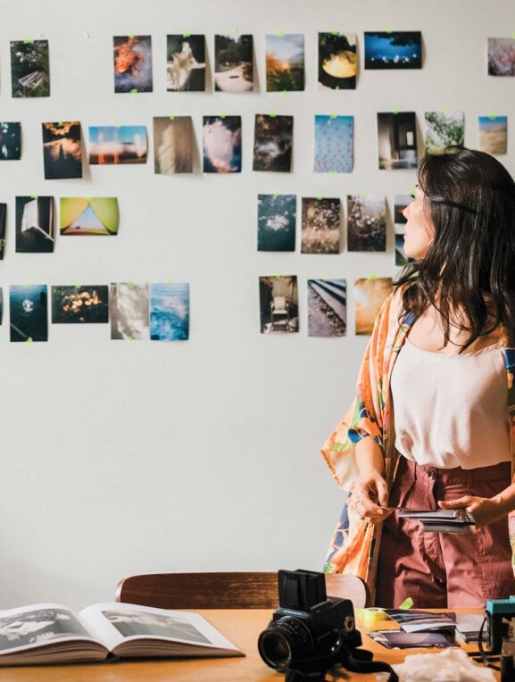 A person stands in front of a wall adorned with a collage of assorted photographs, facing away from the camera. They are surrounded by a creative workspace that includes photography equipment, an open book, a potted plant, and various personal items on a wooden table.