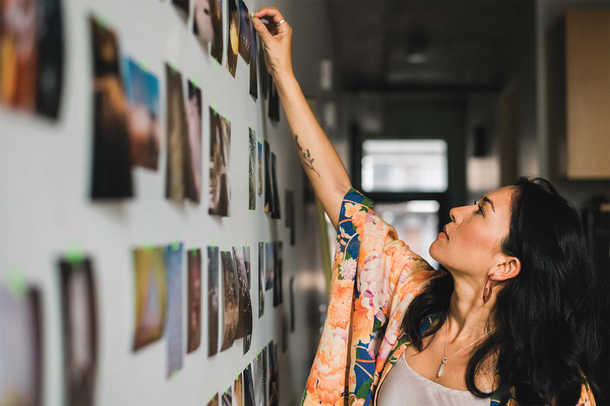 A person with tattoos on the arm wearing a floral print garment reaches out to hang photos on a wall filled with various pictures.
