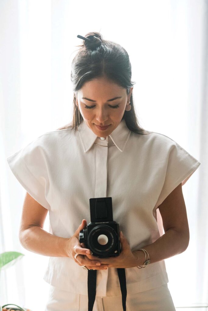A person in a white blouse with a collar, standing in front of a bright window, holding a medium format camera at waist level. The individual has long dark hair styled in a top bun with a black object securing it, and they are wearing a bracelet on their right wrist.