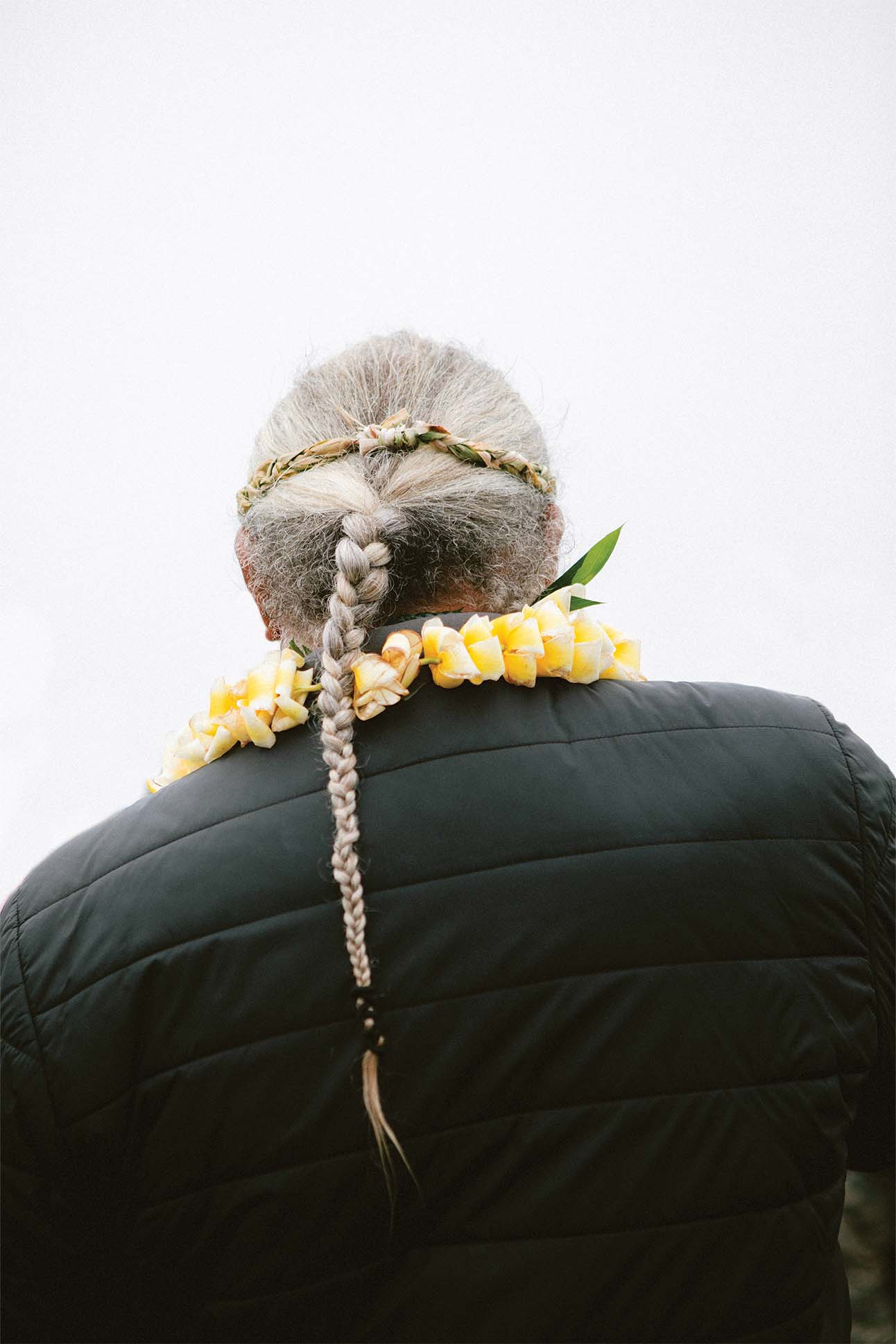 Back view of a person wearing a black jacket with a gray braided hair decorated with a flower crown and a yellow flower lei.