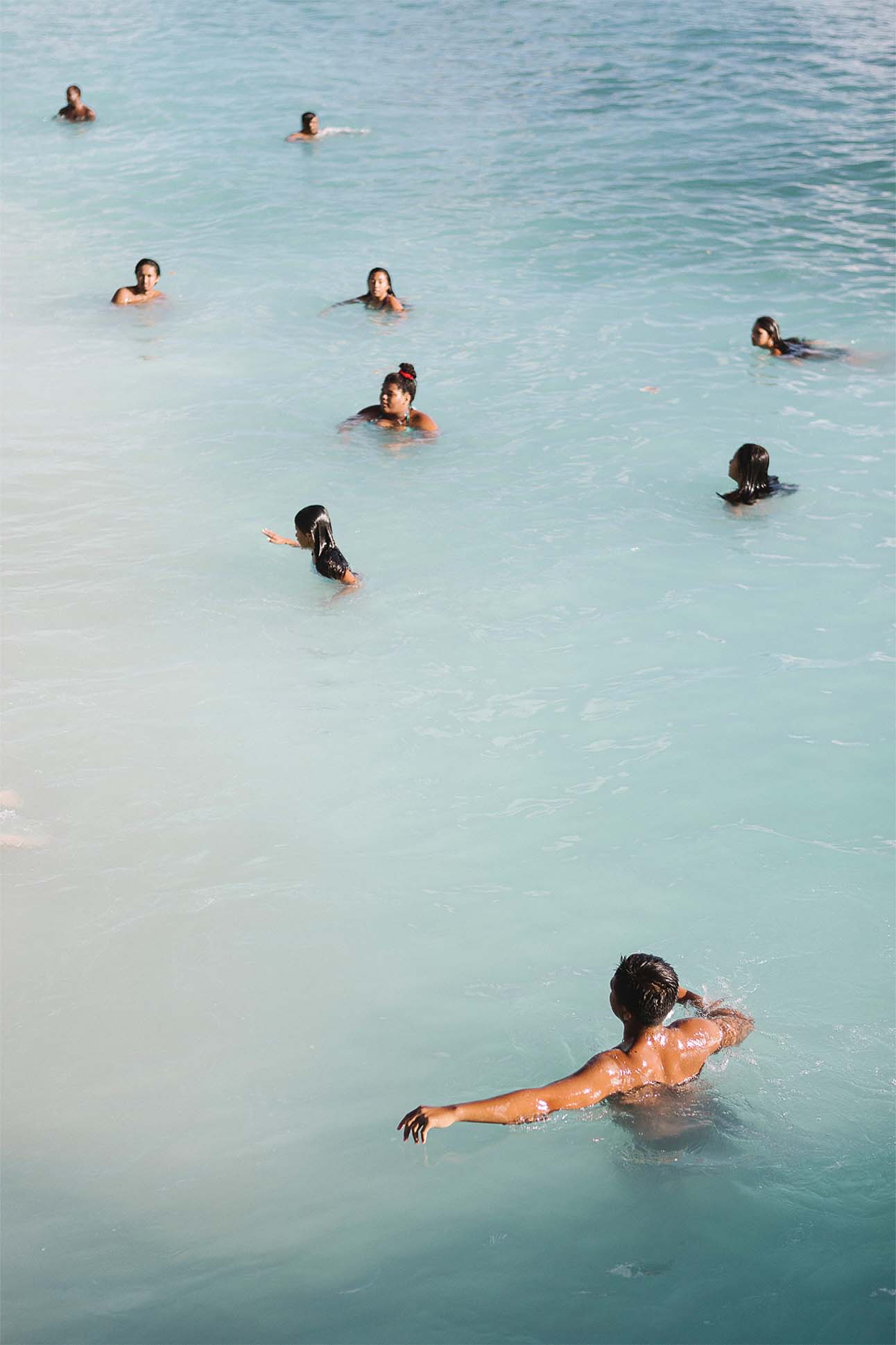People enjoying a swim in clear turquoise waters, with a view of several individuals floating and swimming peacefully. One person in the foreground is seen swimming with arms outstretched gracefully.
