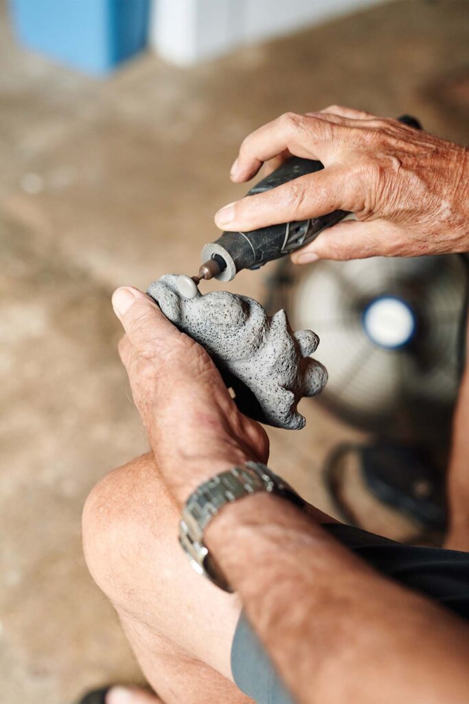 Close-up of a person's hands holding an electric engraving tool and working on a piece of grey stone, with a blurred background.