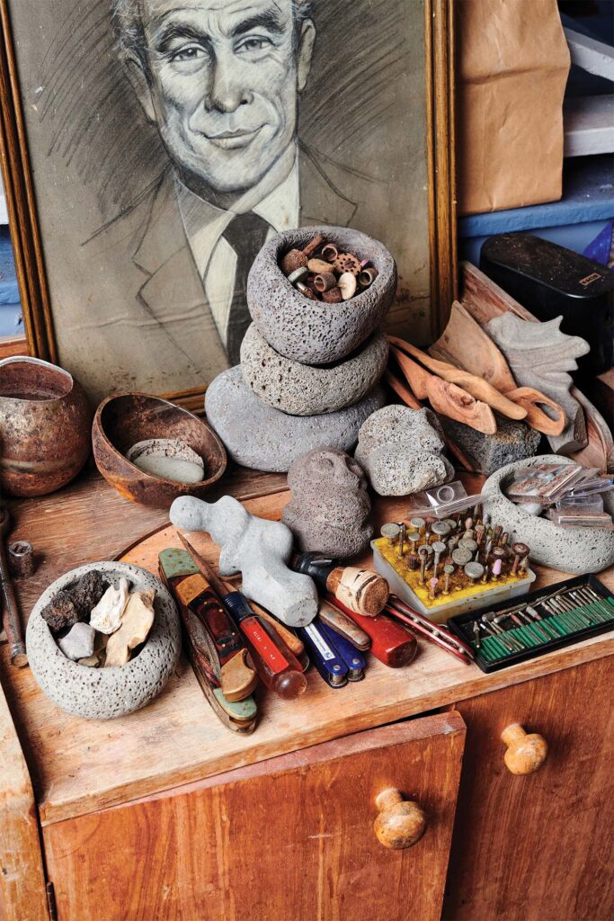 A collection of various items on a wooden work surface, including stone sculptures, wooden bowls, a box of nails, screwdrivers, a wrench, chisels, and other tools, with a partially visible framed artwork propped against the wall.
