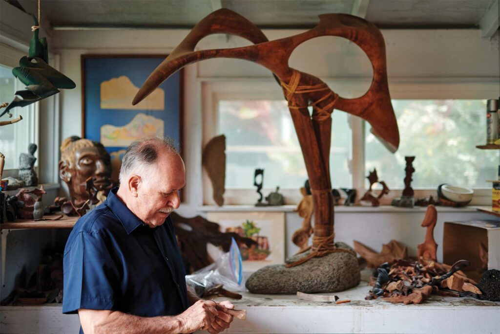 A person sits at a cluttered workbench filled with various wood carving tools and pieces of wood. In the background, a large wooden sculpture resembling a moose's antlers stands prominently, and the room is scattered with many other artistic objects and framed artworks. A window reveals a view of greenery outside.
