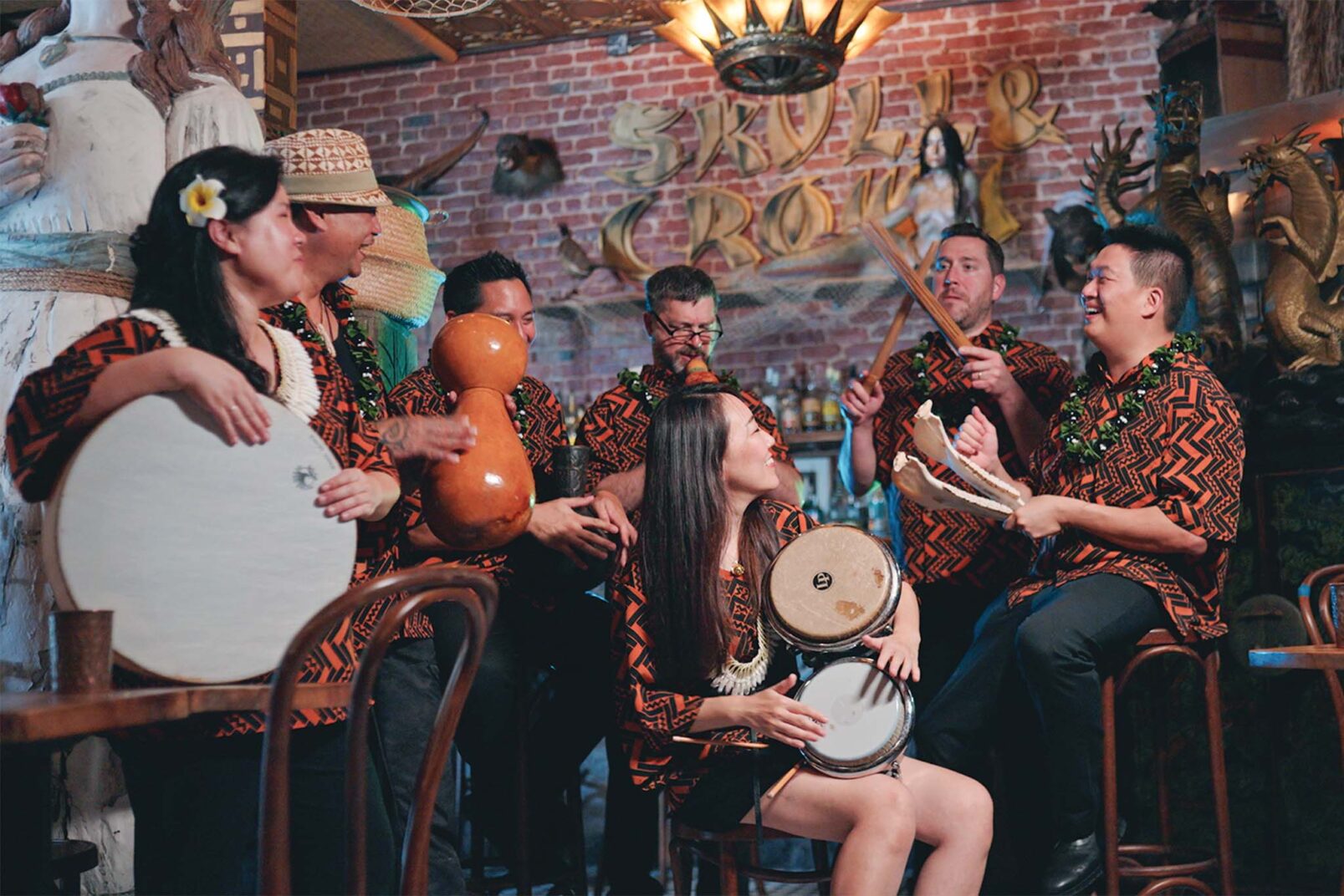 A group of musicians wearing matching traditional patterned shirts is performing with various instruments in a room with eclectic decor, including a sign that reads "SKULL CROWN" and various sculptures on the walls.
