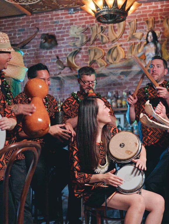 A group of musicians wearing matching traditional patterned shirts is performing with various instruments in a room with eclectic decor, including a sign that reads "SKULL CROWN" and various sculptures on the walls.