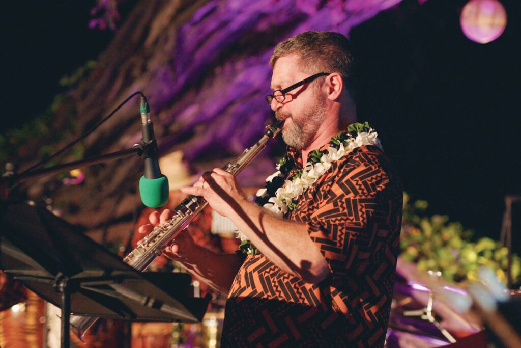 A musician in a patterned shirt with a lei around his neck playing a flute on stage, with music stand in the foreground and ambient lighting in the background.