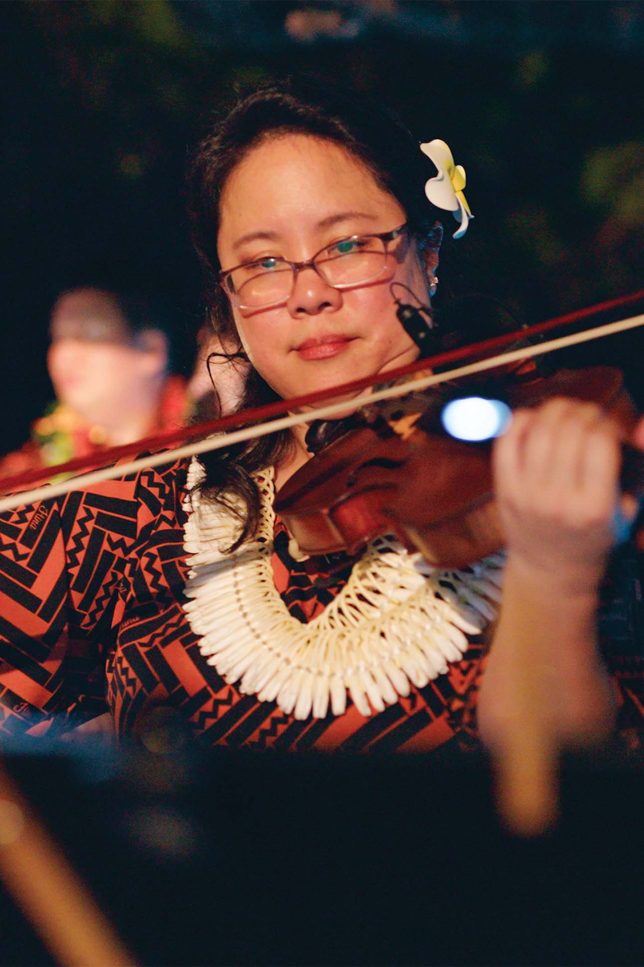 A person wearing a traditional patterned shirt and a shell lei plays the violin at a low-lit evening event, with a white flower tucked behind their ear.