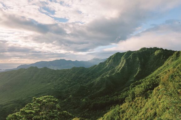 Lush green mountains covered with vegetation under a cloudy sky with sunlight peeking through.