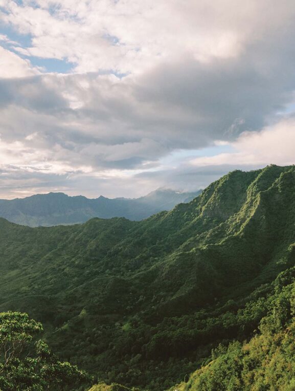 Lush green mountains covered with vegetation under a cloudy sky with sunlight peeking through.